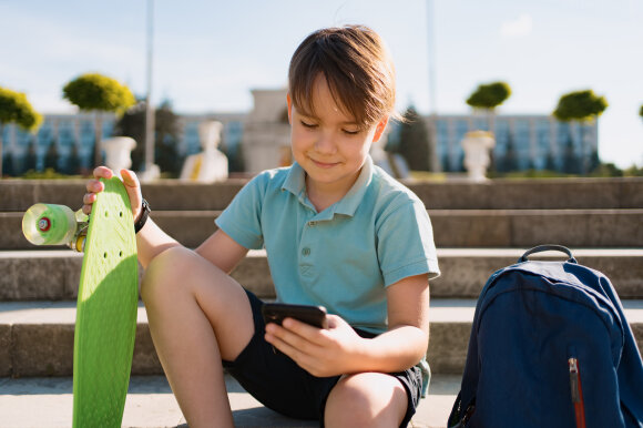 School Boy in blue polo shirt sitting on the stairs with a blue backpack and green penny board using smartphone