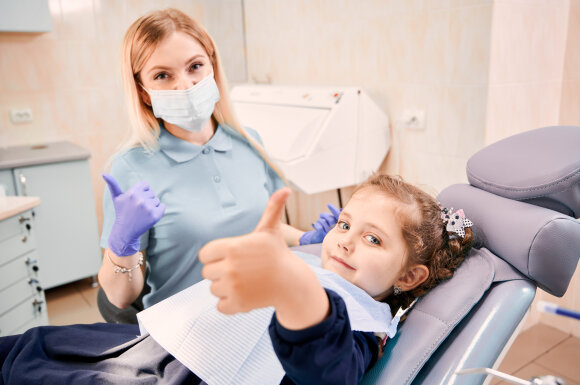 Focus on girl's hand giving thumbs up while woman dentist in medical mask sitting beside kid, showing approval gesture after dental procedure. Concept of pediatric dentistry and dental care approval.